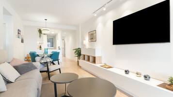 Un salon moderne avec un canapé, deux tables rondes, un téléviseur fixé au mur et une cuisine en arrière-plan.