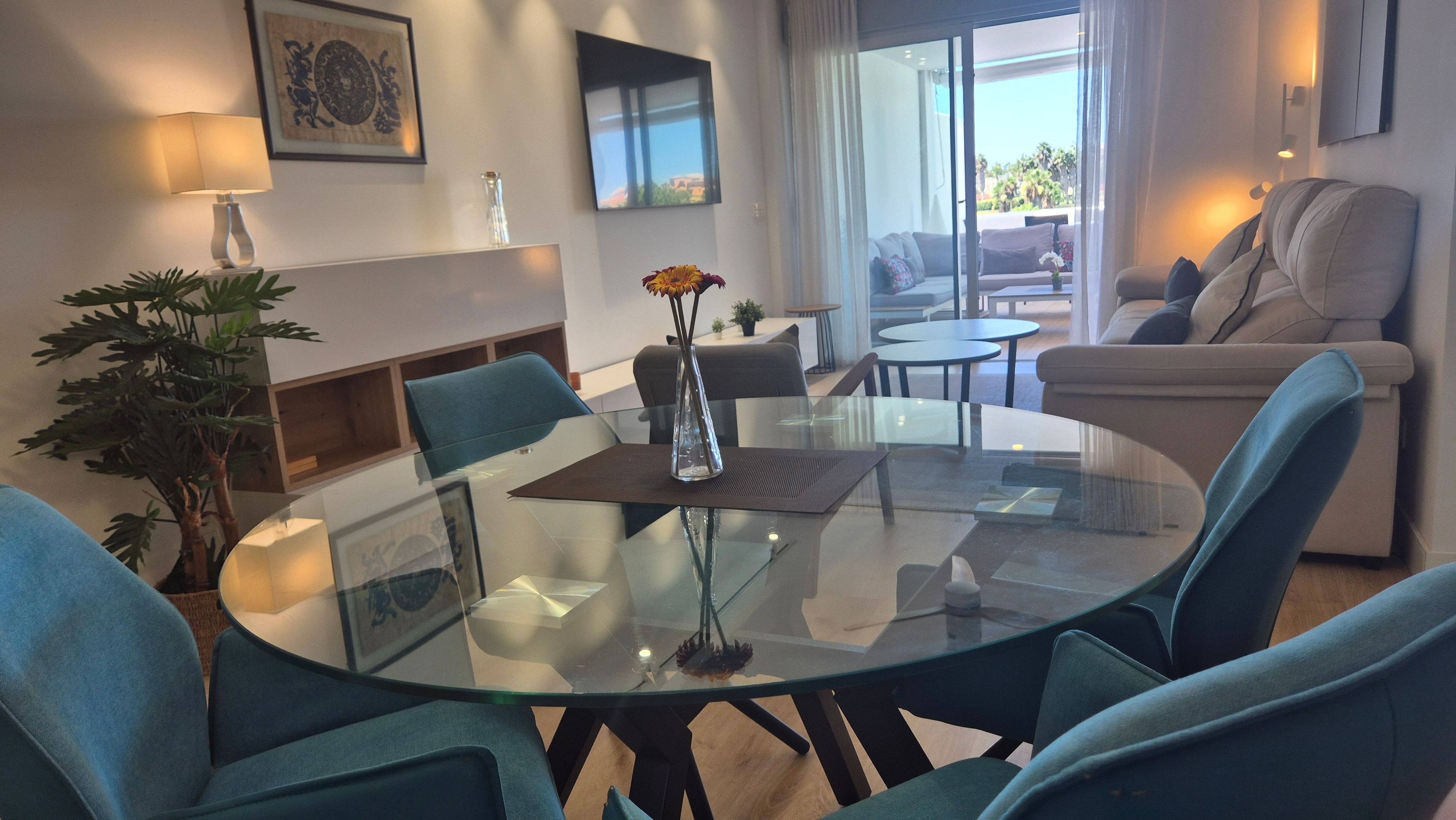 Ein modernes Wohnzimmer mit einem Glastisch, blauen Stühlen, einer Couch und Blick ins Freie.