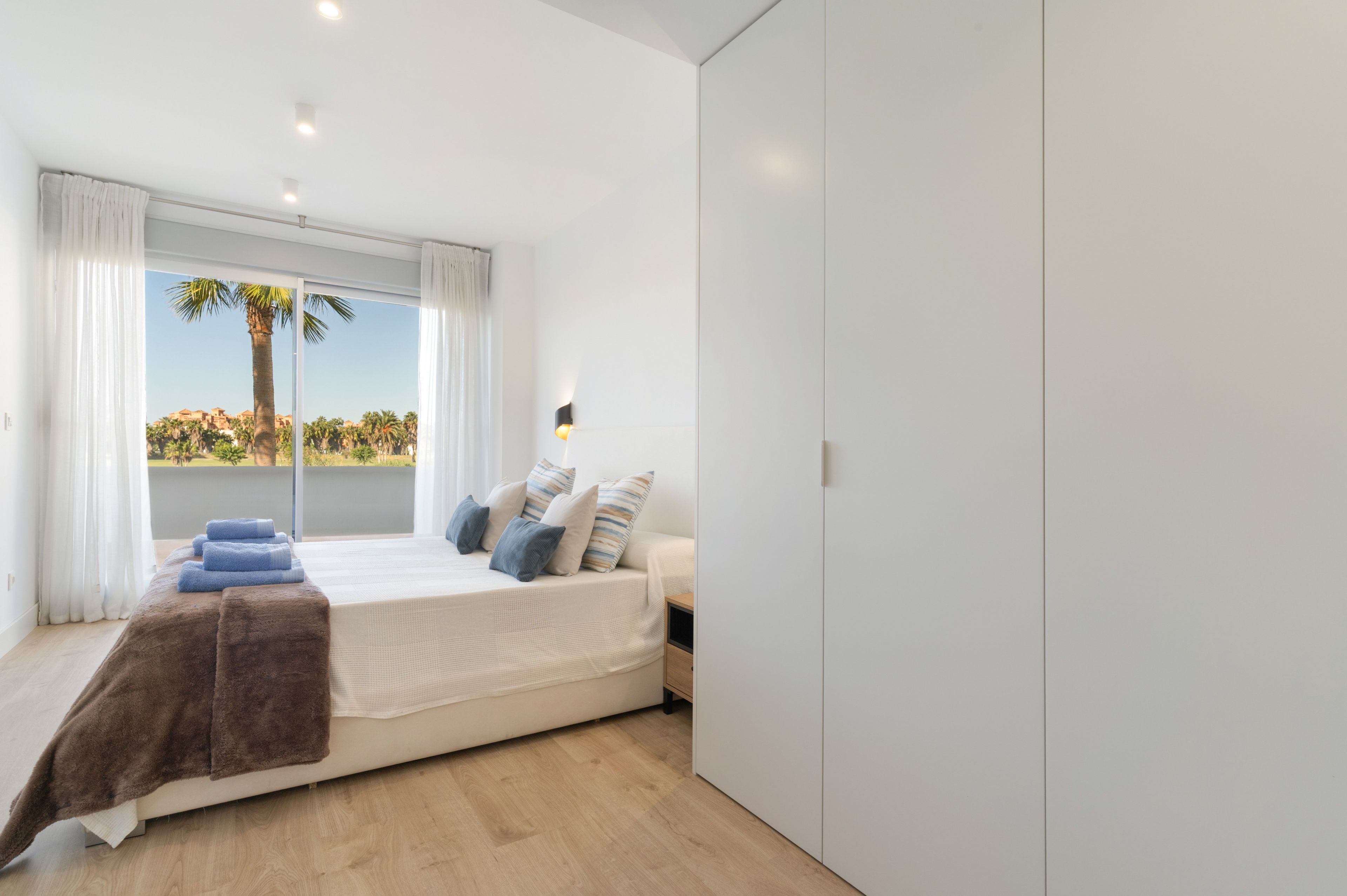 Een slaapkamer met een bed, een raam met gordijnen, een nachtkastje en uitzicht op palmbomen en gebouwen.