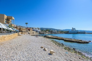A pebbled beach with buildings and a pier, clear skies, and calm water.
