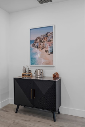 A black cabinet with a geometric pattern, a framed coastal landscape painting, and a small wooden tray with kitchen items. - The Cabana Club-3BR- Fantastic Outdoor Pool-Bar-TV (Fort Lauderdale)