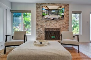 A modern living room with a stone fireplace, two armchairs, a coffee table, and large windows with a view of greenery. - The Cabana Club-3BR- Fantastic Outdoor Pool-Bar-TV (Fort Lauderdale)