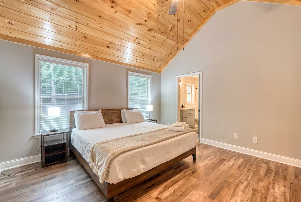 Camera da letto con soffitto in legno, un letto grande, due finestre e bagno visibile attraverso una porta aperta.