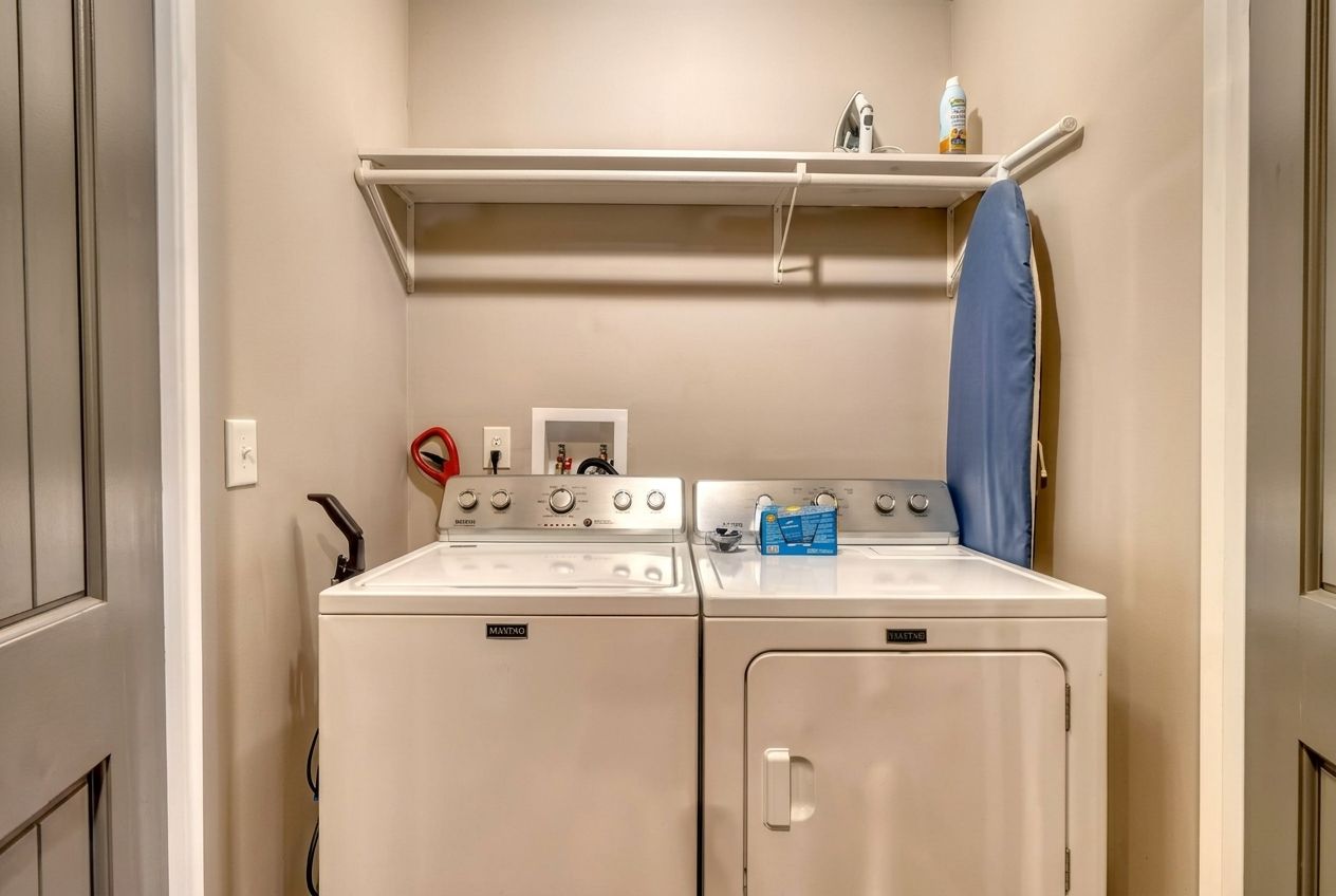 A compact laundry area with two washing machines, a shelf above, and a blue curtain.