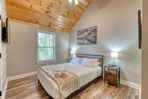 Une chambre avec un plafond en bois, un lit avec une literie blanche, une fenêtre avec des stores, une table de chevet avec une lampe et un téléviseur fixé au mur.