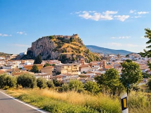 A coastal town with a prominent hilltop fortress, a road with a directional sign, and residential buildings. - New Listing (Íllora)