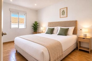 Un lit bien fait, avec une tête de lit, deux tables de chevet avec des lampes, une plante en pot et une fenêtre donnant sur un immeuble.