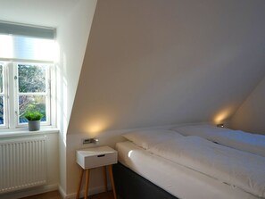 A bedroom with a window, a bedside table with a drawer, and a bed with white bedding. - 6 St. Barth in the Boldix House (Wyk auf Foehr)