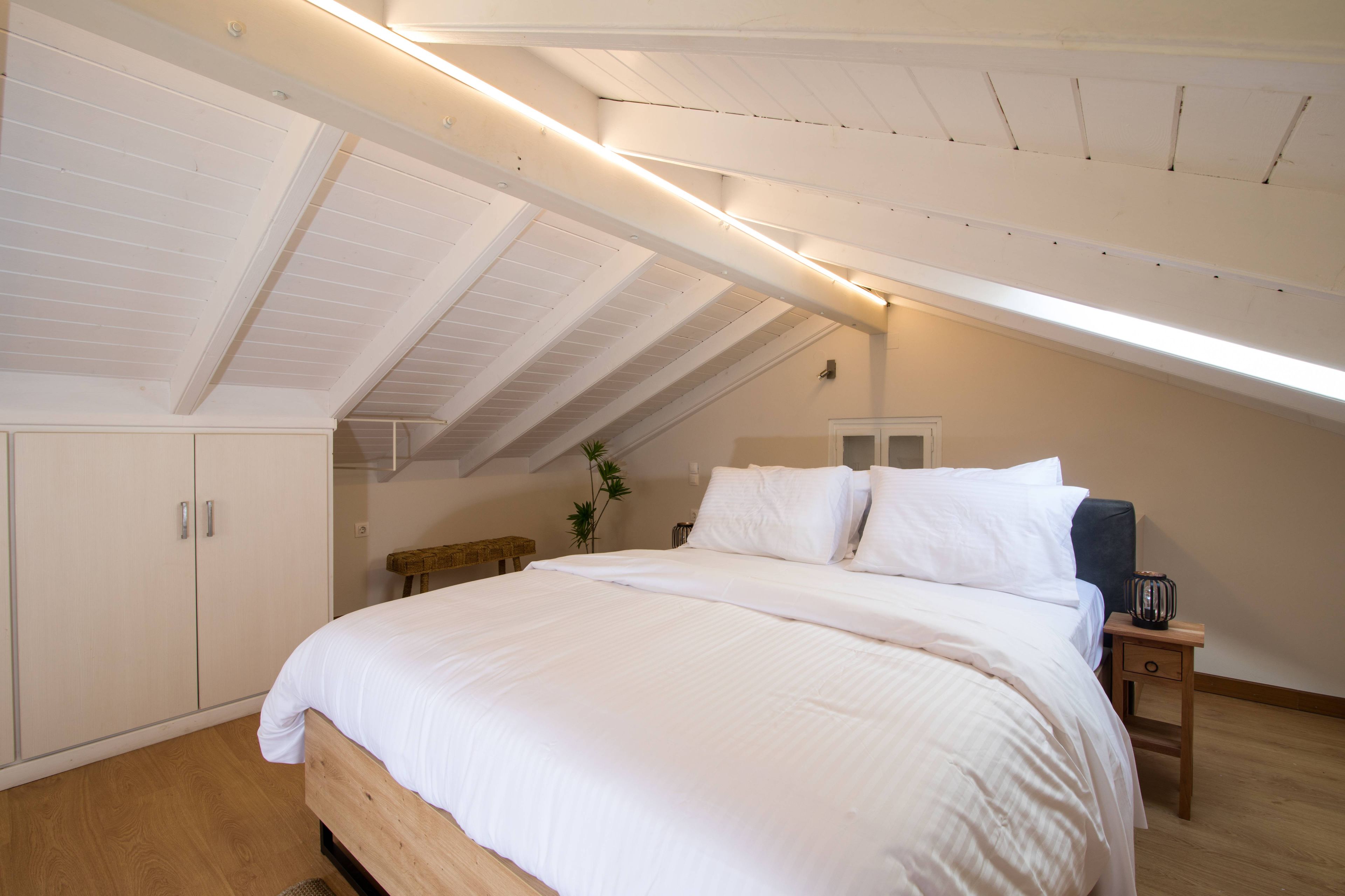 A bedroom with a sloped ceiling, a bed with white bedding, a bedside table with a lamp, and a small plant.