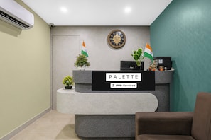 Mostrador de recepción con un cartel que dice 'PALETTE' y 'OYO-Serviced'. Hay dos plantas en macetas y dos banderas en la pared detrás del mostrador.