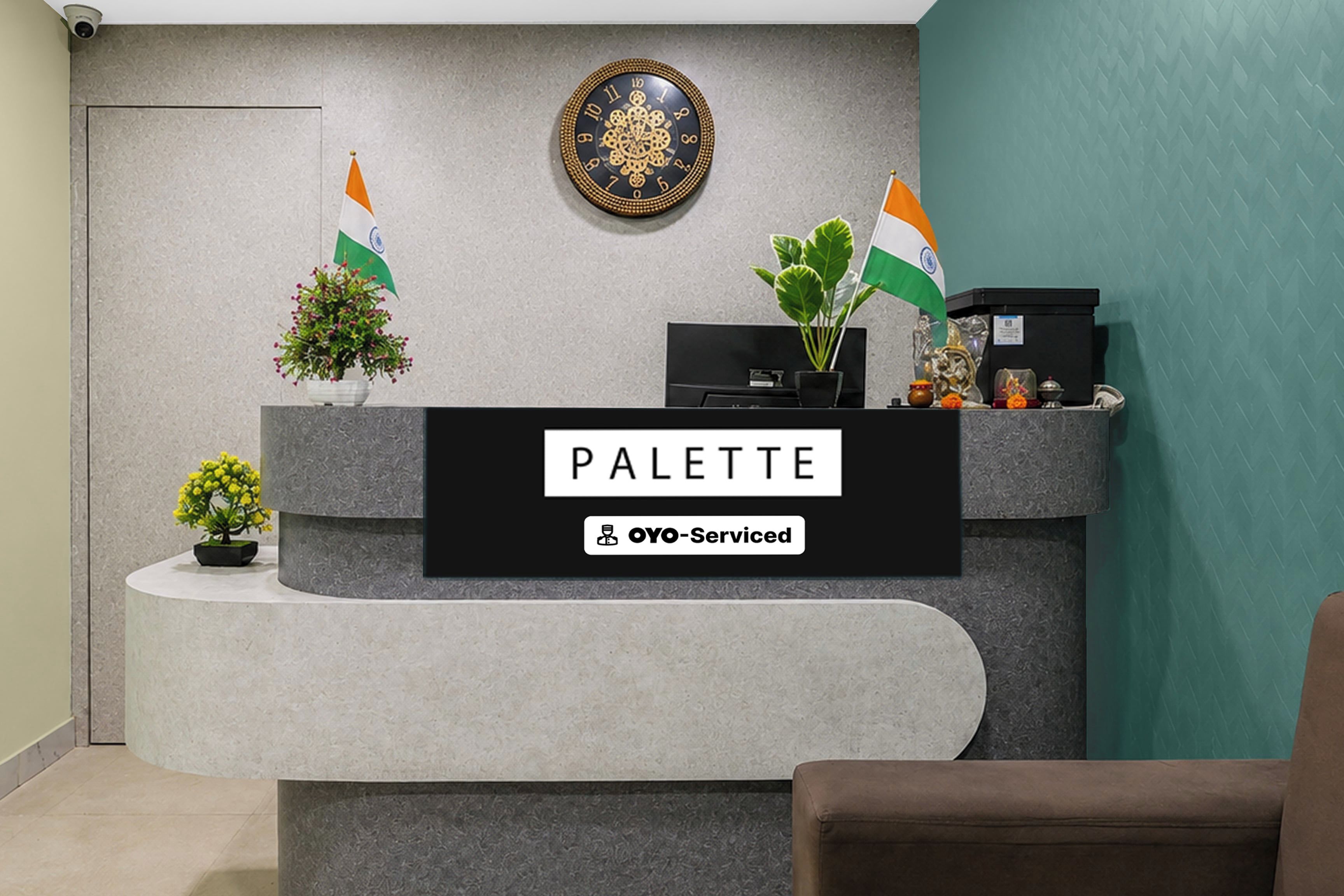 Un mostrador de recepción con un letrero que dice 'PALETTE' y 'OYO-Serviced'. Hay dos banderas, una planta en maceta y una pequeña estatua sobre el mostrador.