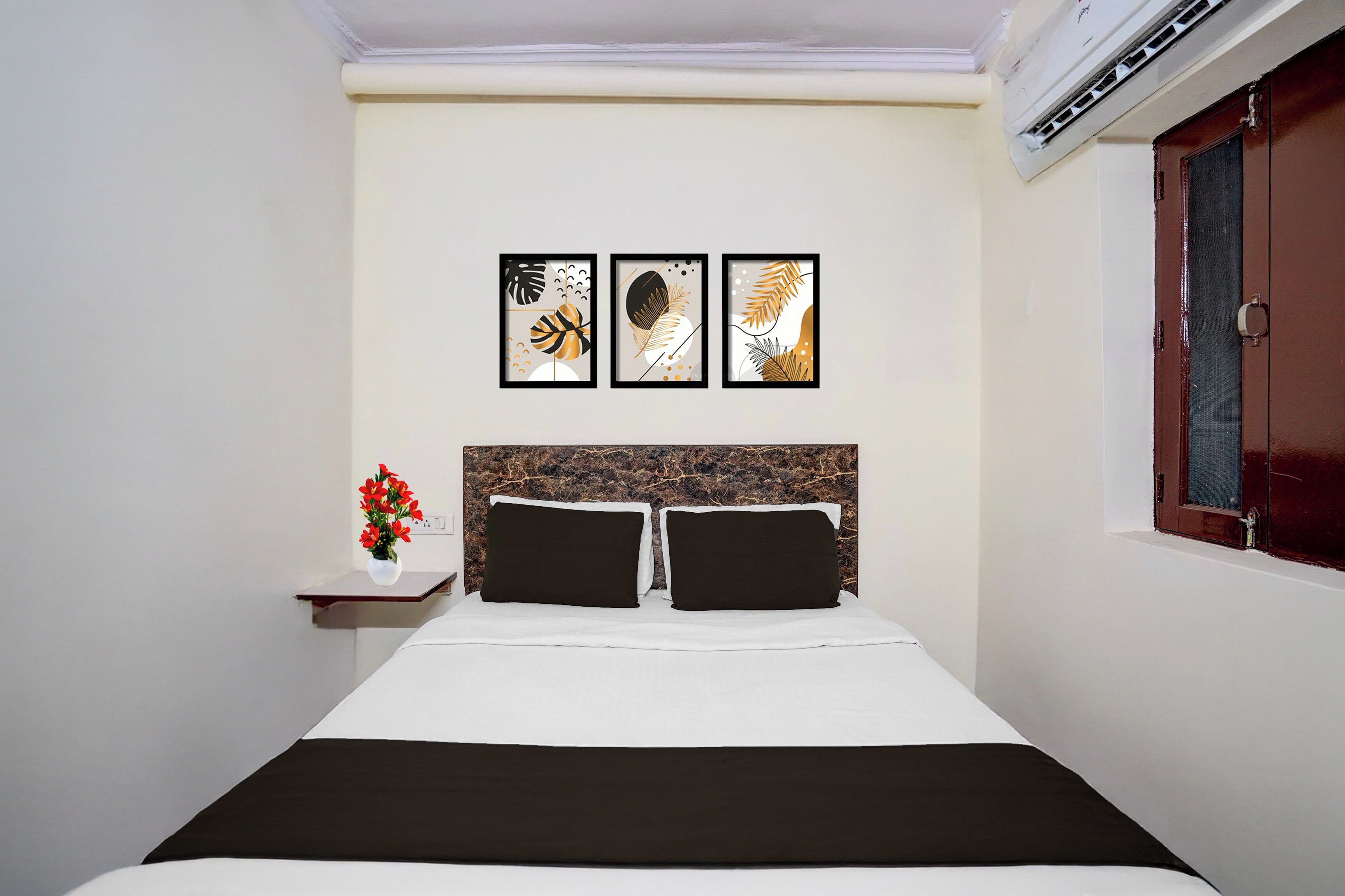 A hotel room with a bed, a nightstand with a vase of flowers, and three framed artworks on the wall.