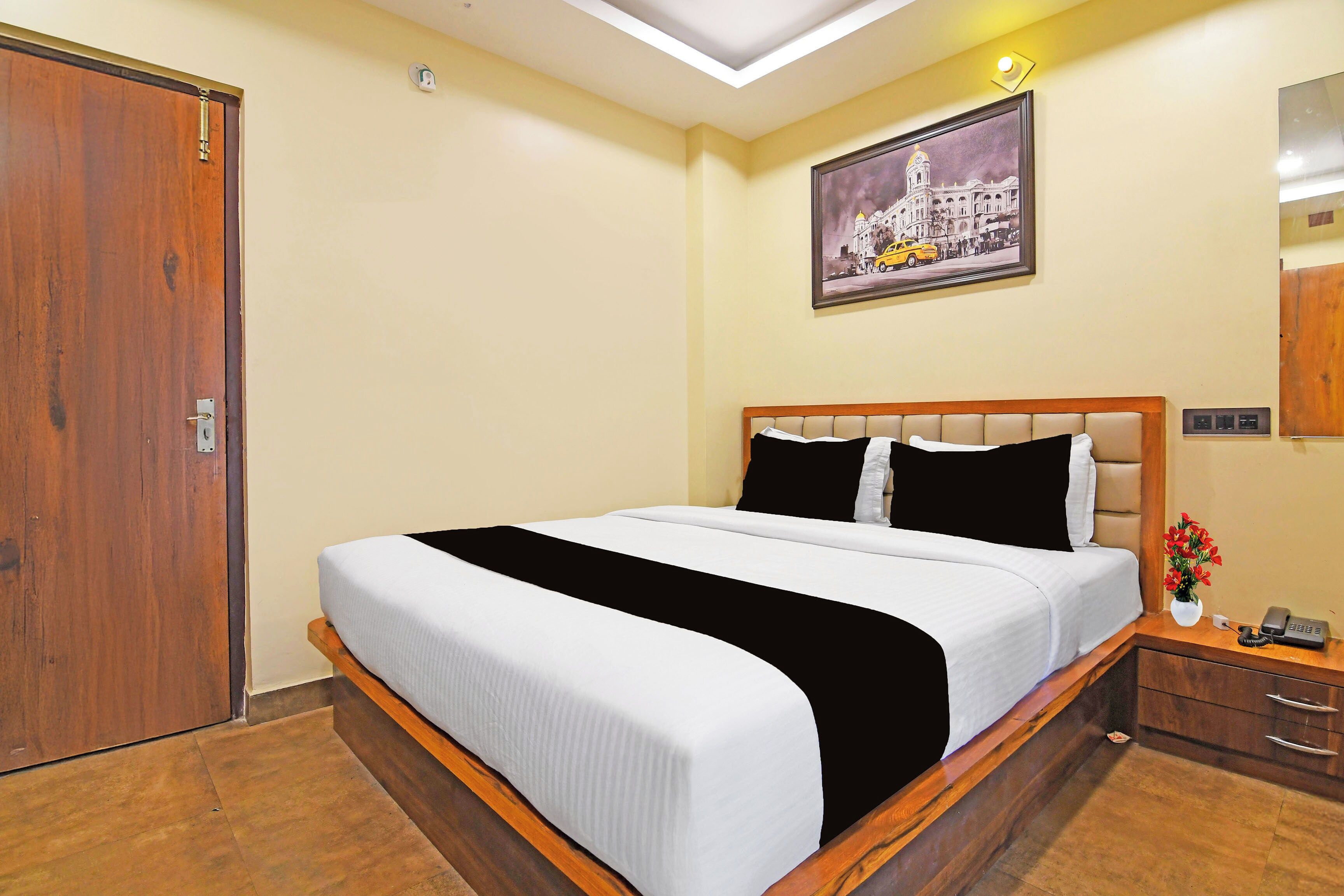 A hotel room with a large bed, a wooden headboard, a nightstand with a phone, a wall-mounted light, and a painting of a building.