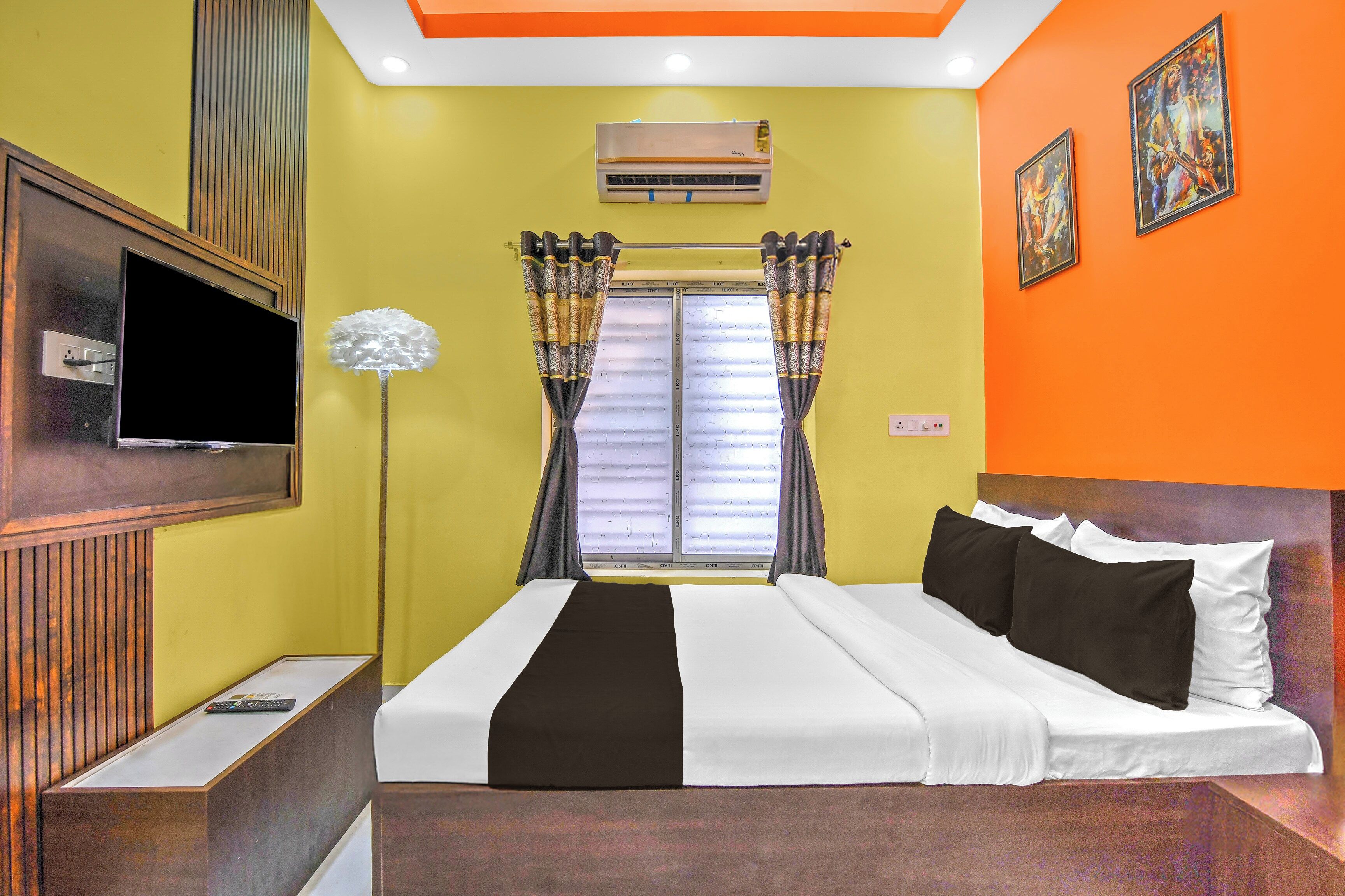 A hotel room with a bed, a TV, an air conditioning unit, a window with curtains, and a painting on the wall.