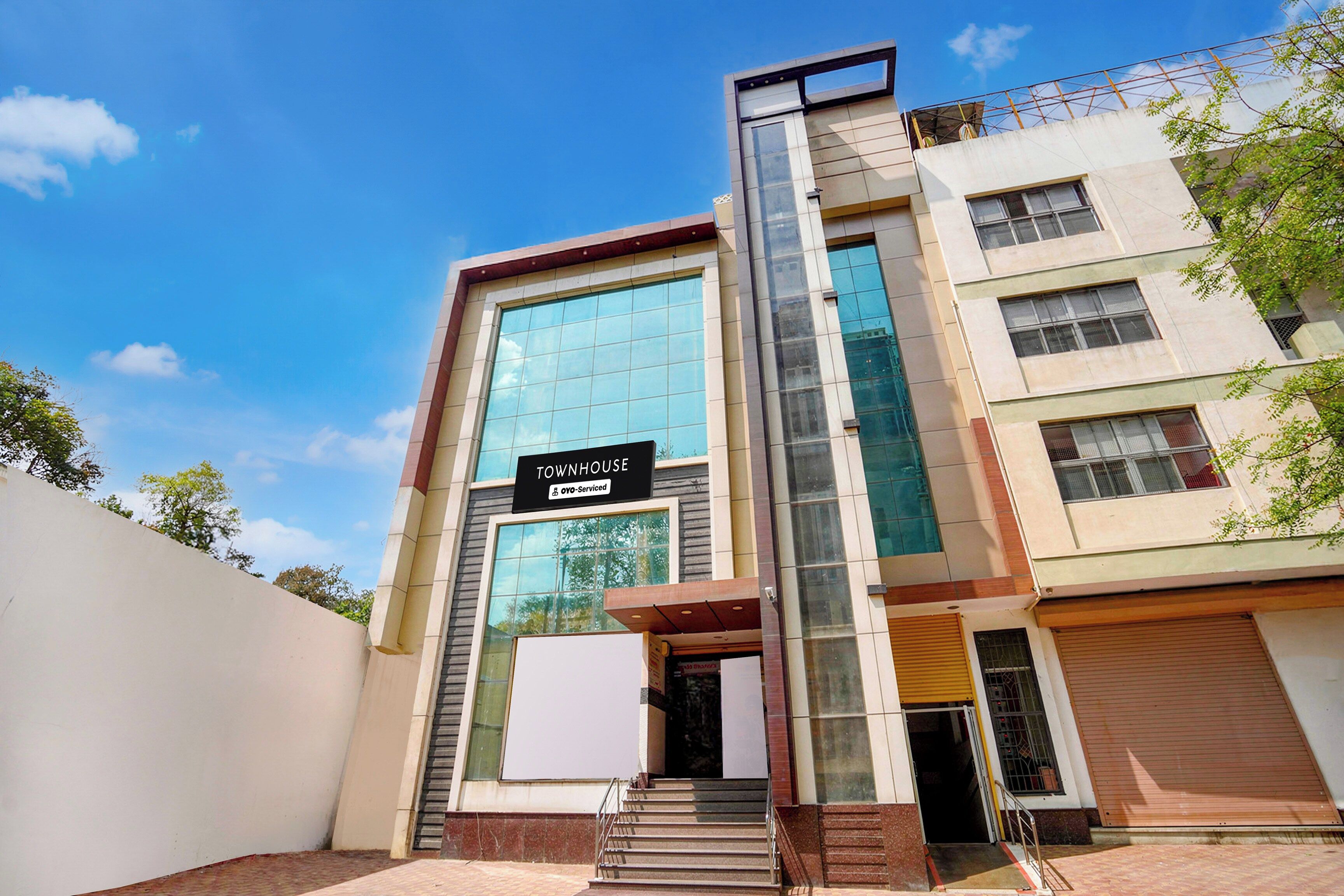 A modern townhouse with a prominent glass facade, a sign indicating it is serviced by OYO, and a set of stairs leading to the entrance.