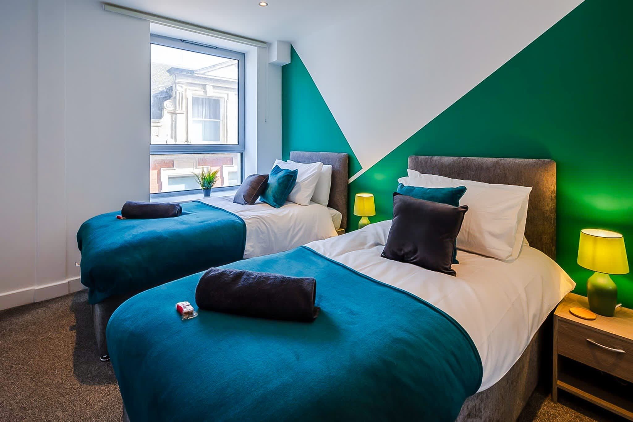 A hotel room with two beds, a green accent wall, a window with a view, and a bedside table with a lamp.