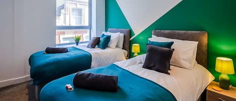 A hotel room with two beds, a green accent wall, a window with a view, and a bedside table with a lamp.