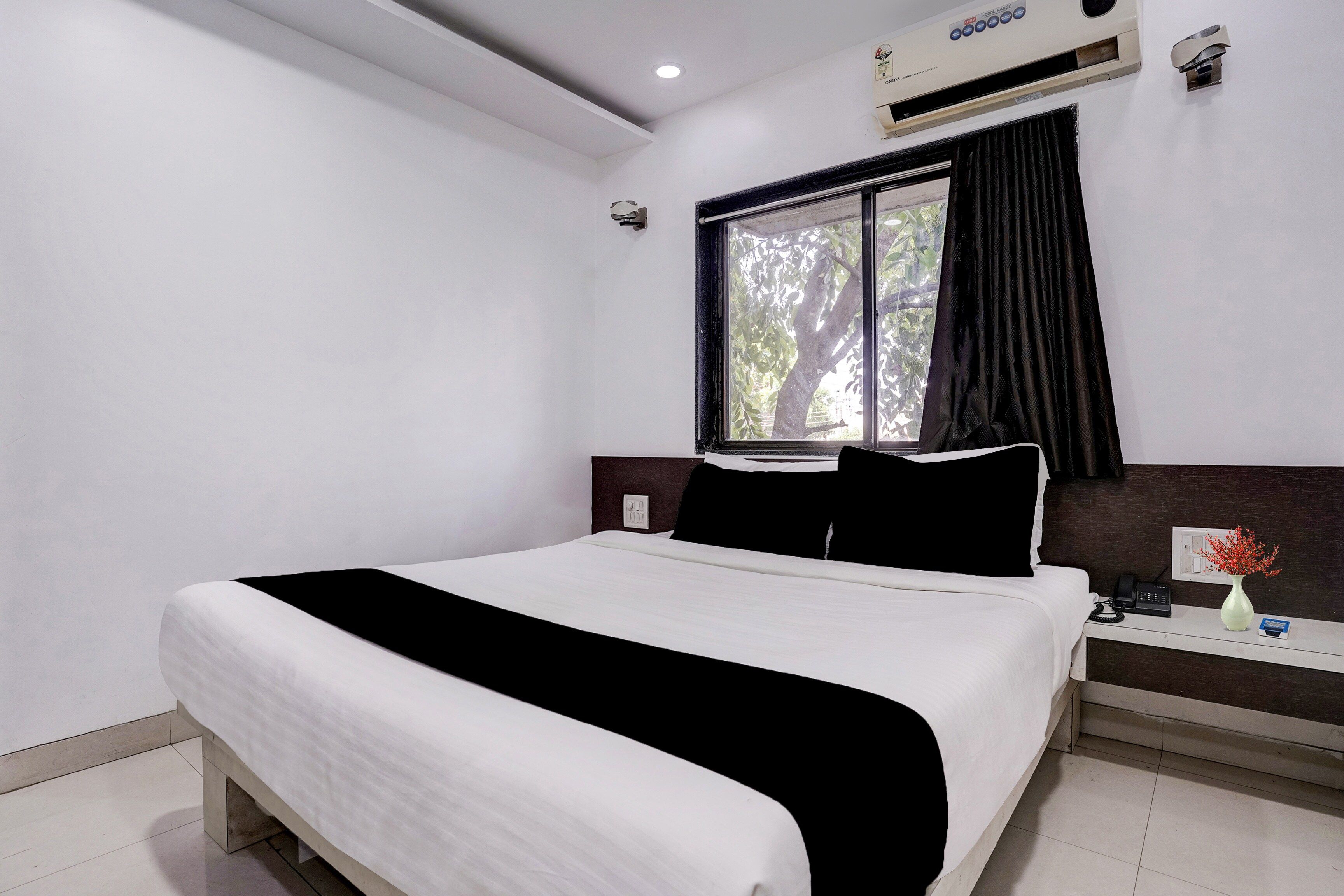 Una habitación de hotel con cama, mesita de noche, un jarrón con flores y una ventana con cortinas.
