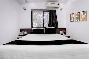 Une chambre d’hôtel avec un lit, une fenêtre avec des rideaux, un appareil de climatisation, une table de chevet avec une bouilloire et des œuvres d’art encadrées accrochées au mur.
