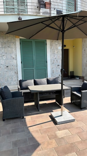 An outdoor seating area with a table and chairs, a large umbrella, and a building with a green shutter in the background. - Apartment 'Casa Mimma' with Private Terrace, Wi-Fi and Air Conditioning (Terni)