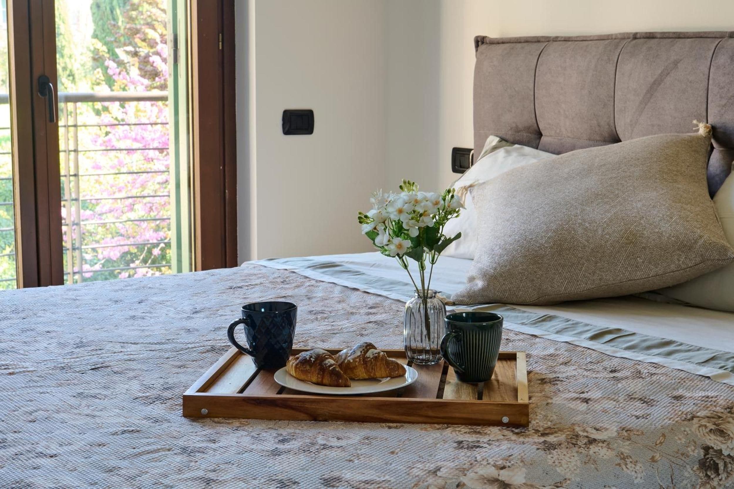 Un lit bien fait, agrémenté d’un plateau de croissants, d’un vase rempli de fleurs et de deux tasses posées sur une couverture au motif texturé.