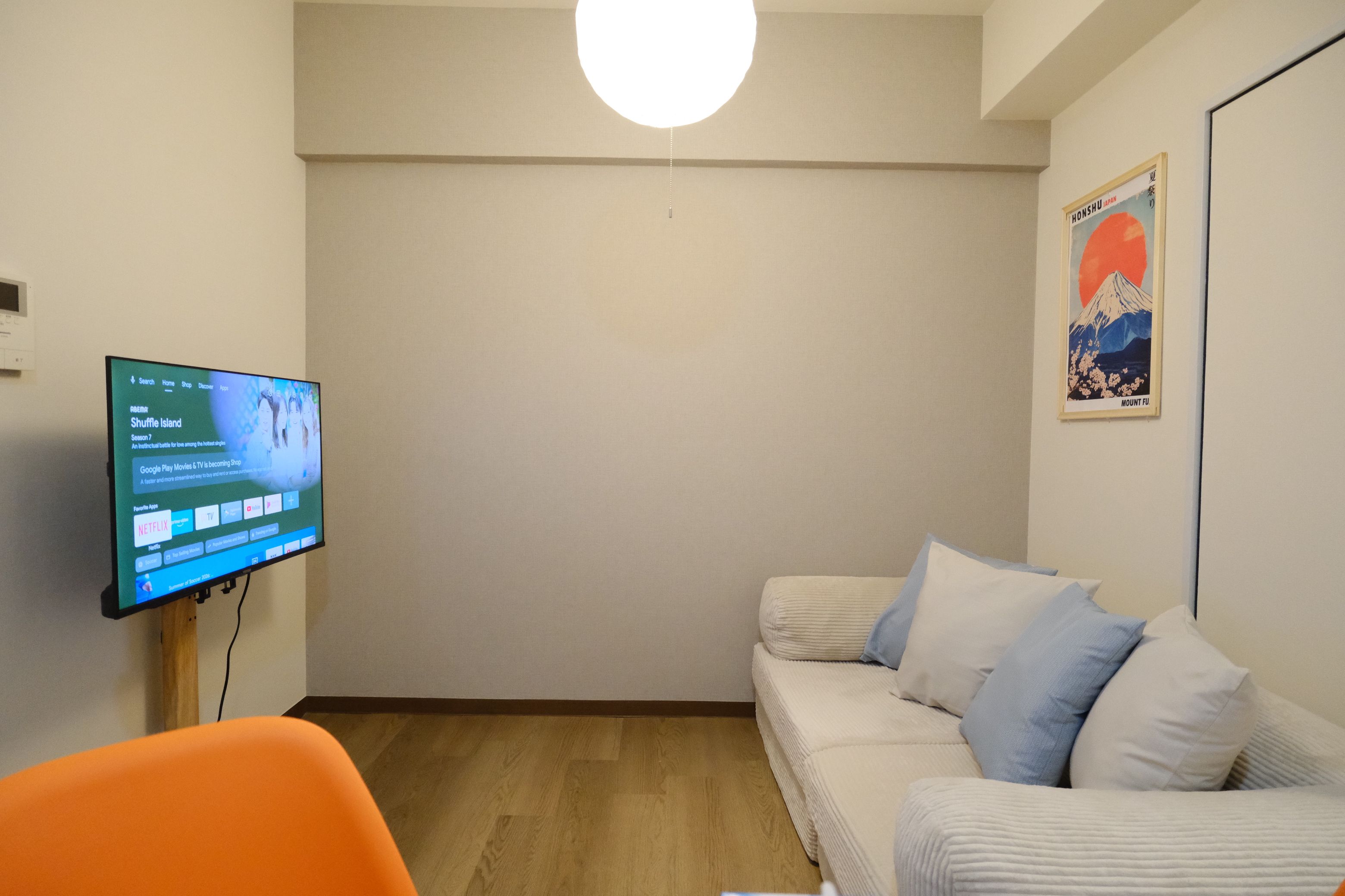 A room with a flat-screen TV mounted on the wall, a white sofa with blue cushions, and a framed picture of Mount Fuji.