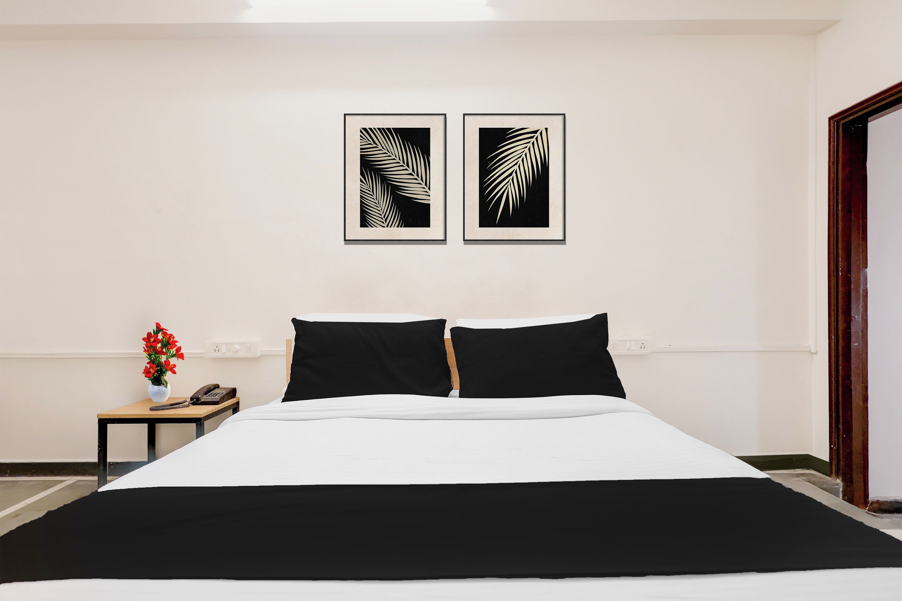 Habitación de hotel con una cama, dos almohadas negras, una mesita de noche con un jarrón de flores rojas y dos cuadros en la pared.