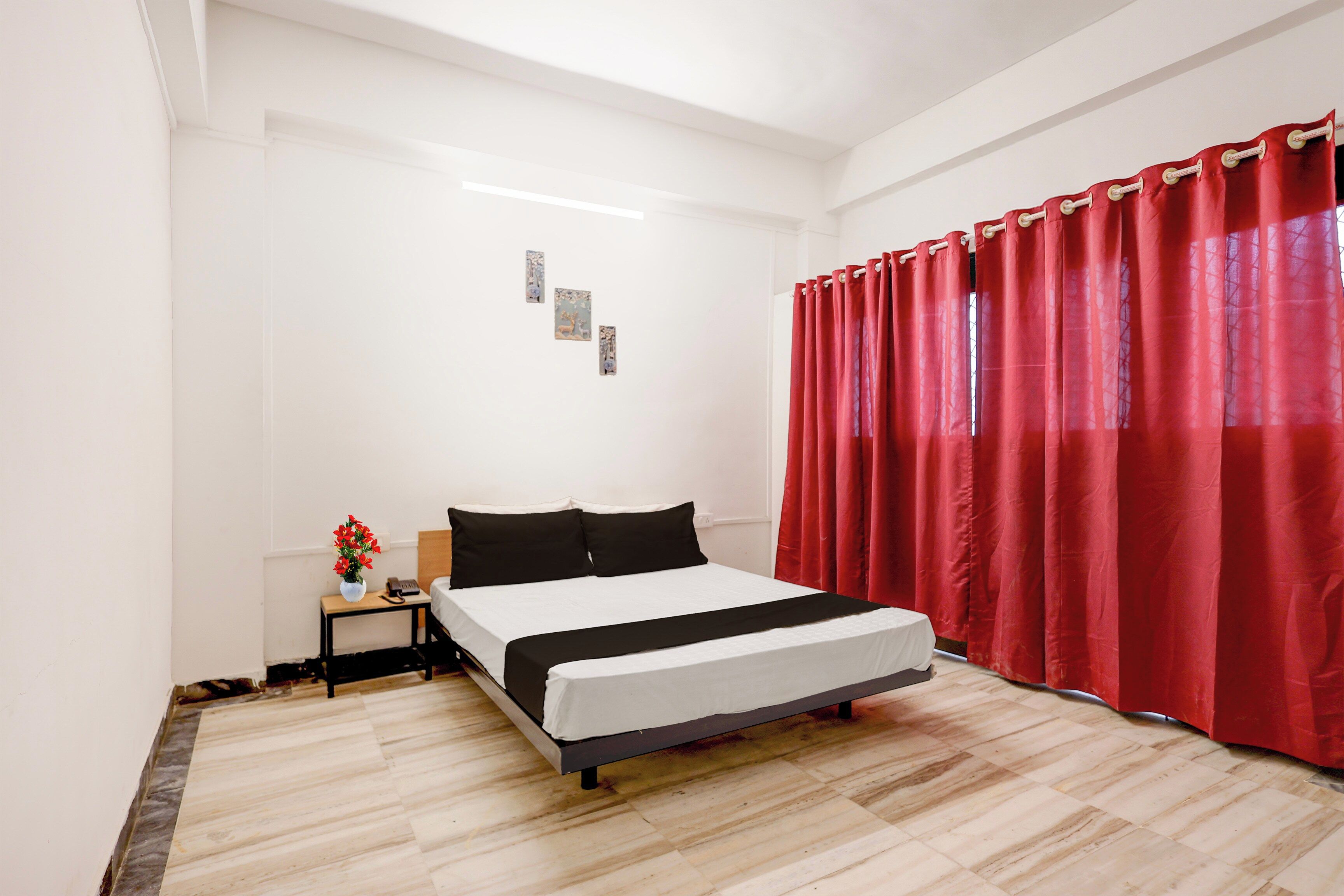 Een slaapkamer met een bed, een nachtkastje, een vaas met bloemen en een rood gordijn.