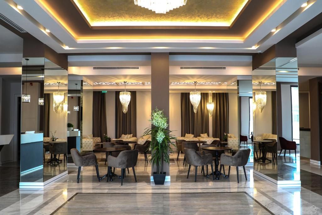 Un vestíbulo de hotel con un mostrador de recepción central, varias zonas de comedor con mesas y sillas, y una gran lámpara de araña.