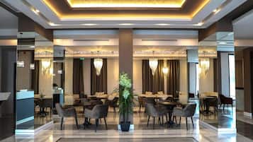 Un vestíbulo de hotel con un mostrador de recepción central, varias zonas de comedor con mesas y sillas, y una gran lámpara de araña.