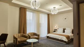 Een hotelkamer met een bed, twee fauteuils, een tafeltje, een klok aan de muur en een raam met gordijnen.