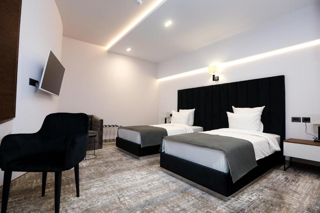 A hotel room with two beds, a black chair, a flat-screen TV mounted on the wall, and a modern light fixture.