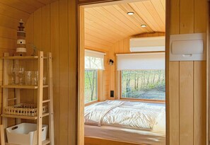 Ein Zimmer aus Holz mit einem Bett, Regalen mit Gläsern und Blick auf Bäume durch das Fenster.