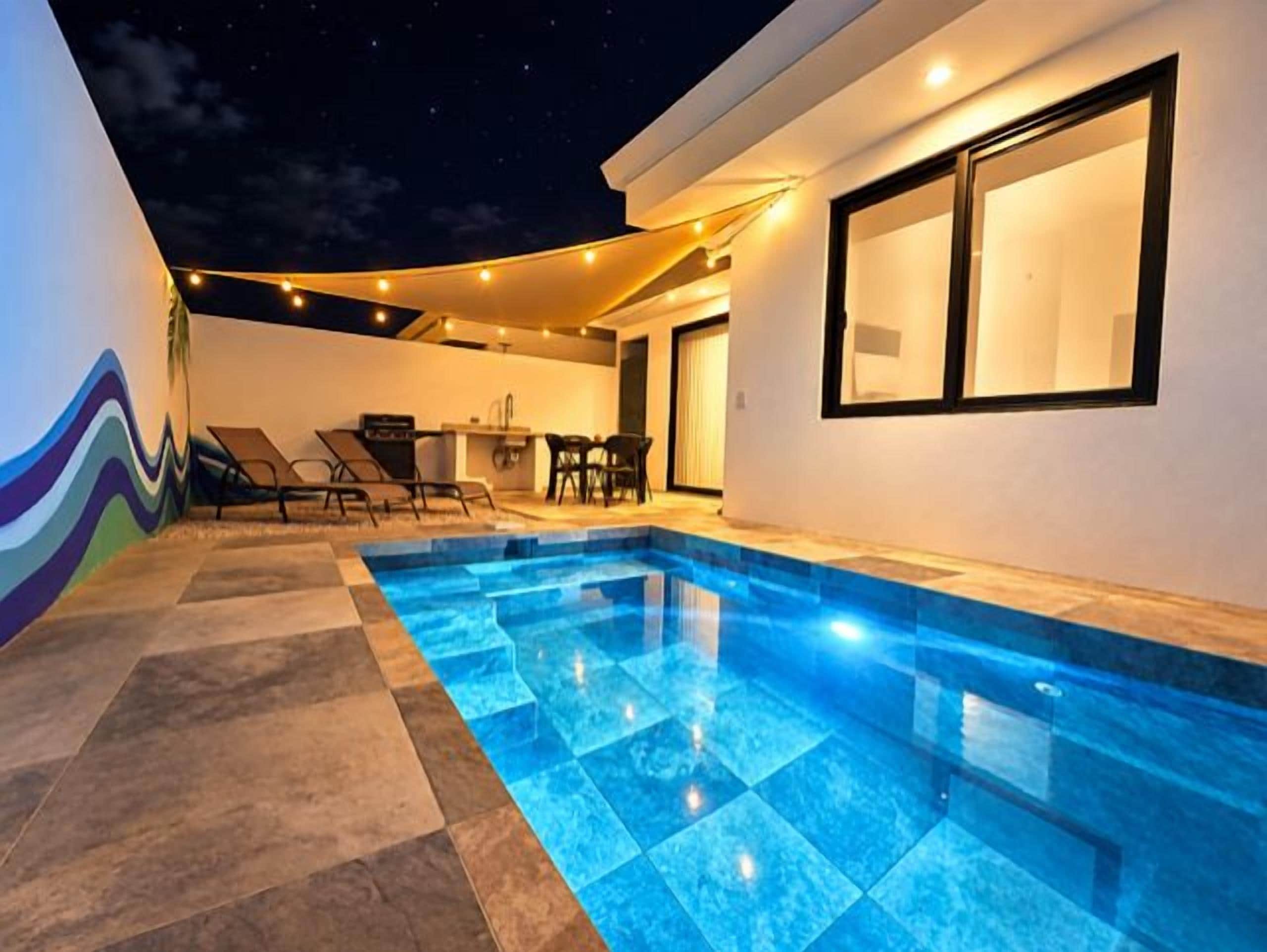 A swimming pool with a tiled deck, a dining area with chairs, and a wall with a wavy design.