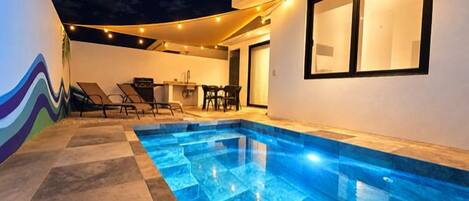 A swimming pool with a tiled deck, a dining area with chairs, and a wall with a wavy design.