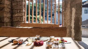 A wooden table with food items and a cup, set against a stone wall and wooden beams, with a view of buildings through a barred window.