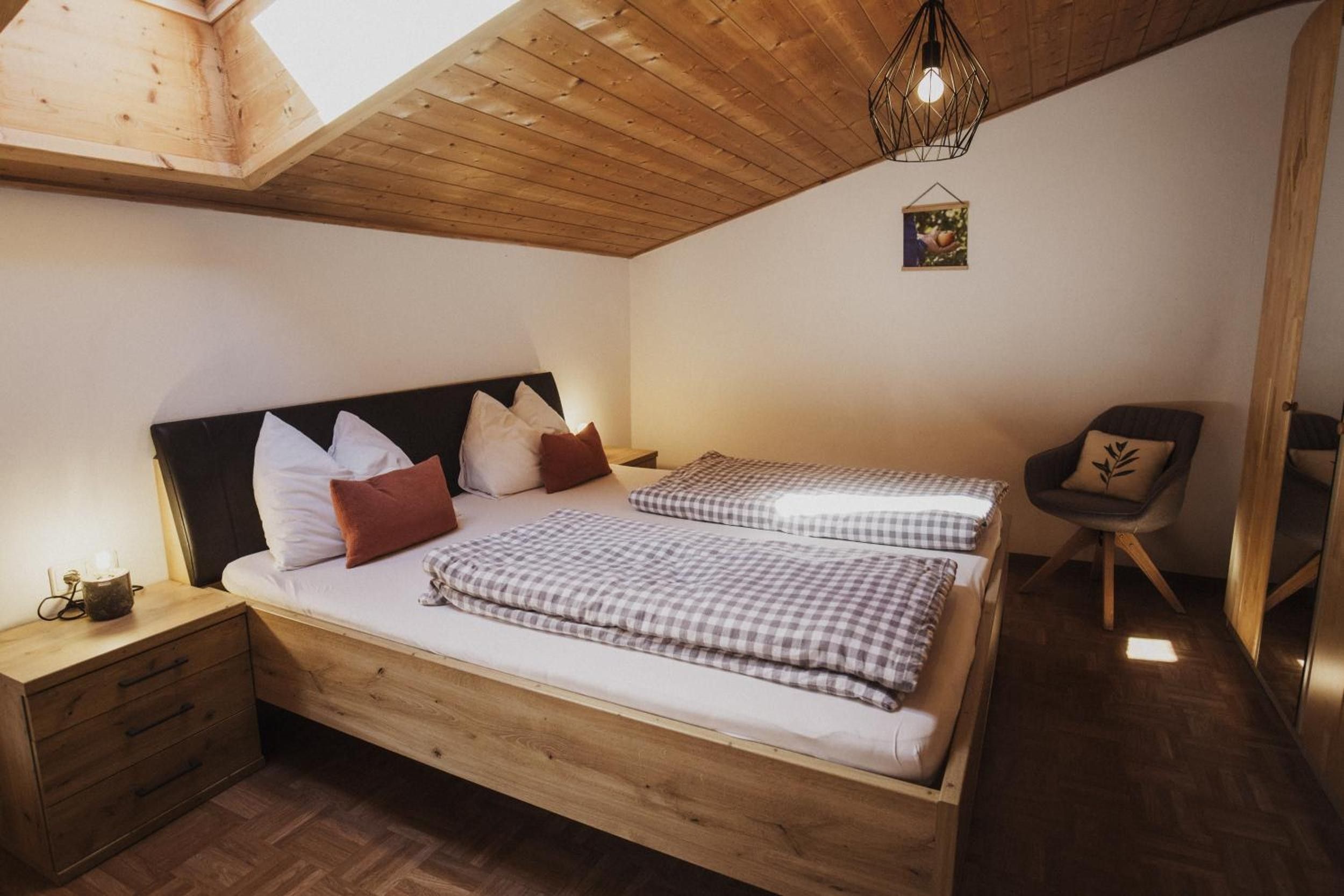 A bedroom with a wooden bed, two pillows, a bedside table, a chair, and a hanging light fixture.