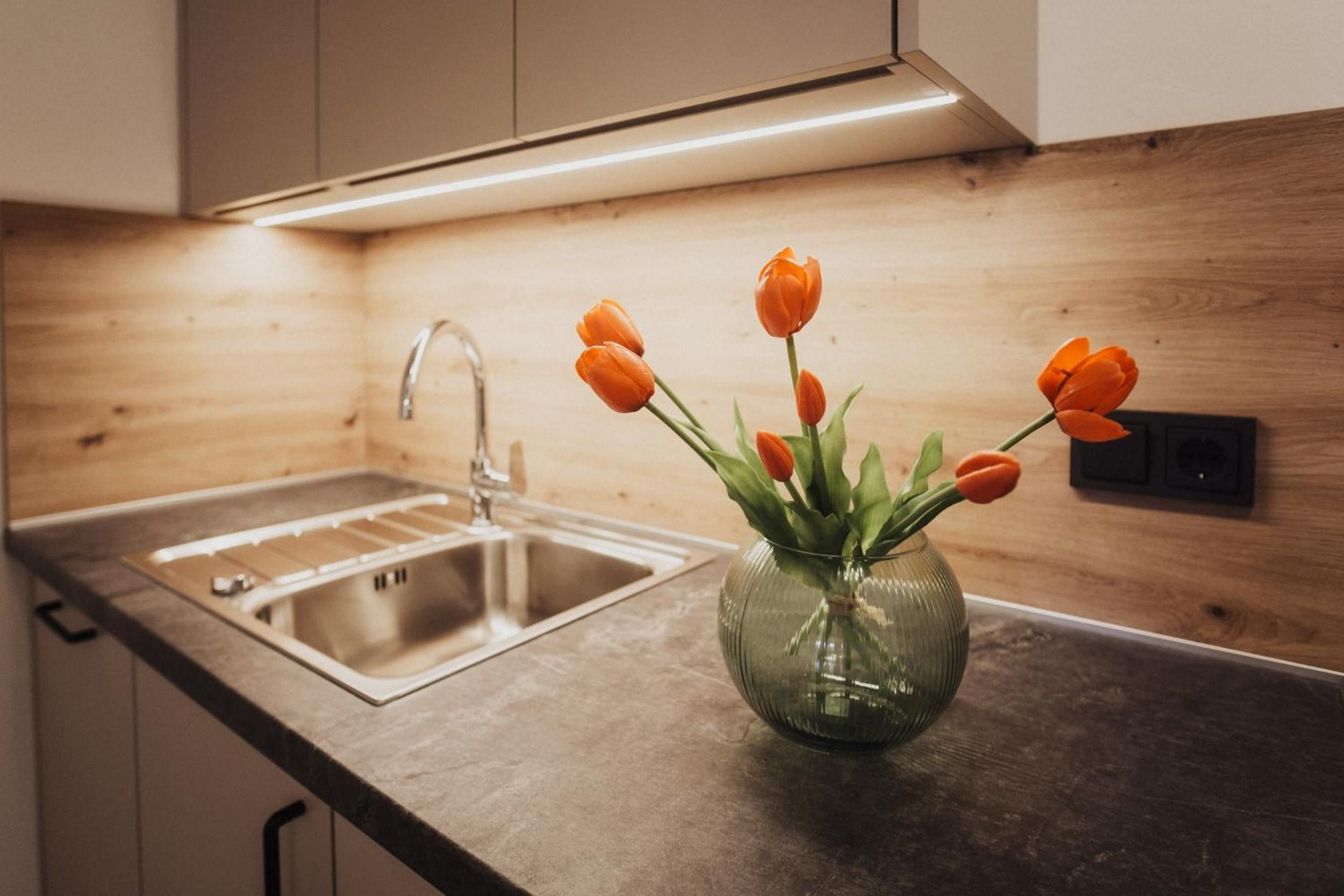 Una cocina con un lavamanos de acero inoxidable, una encimera de madera y un florero de vidrio con tulipanes naranjas.