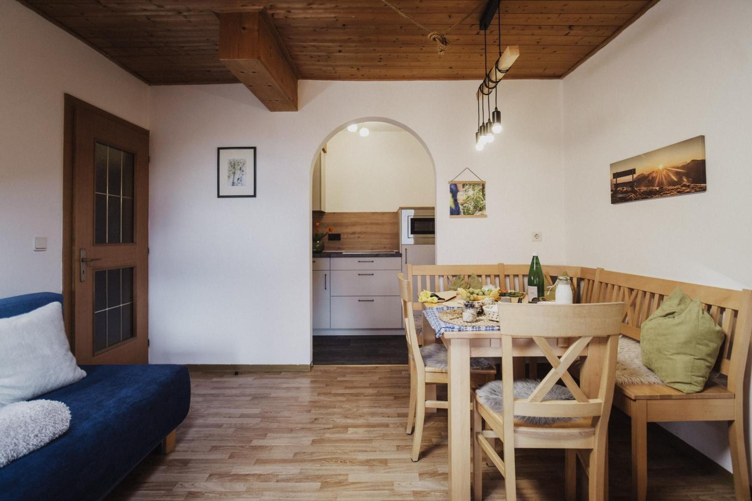 A cozy room with a wooden ceiling, a dining table with chairs, a blue sofa, and a kitchen in the background.