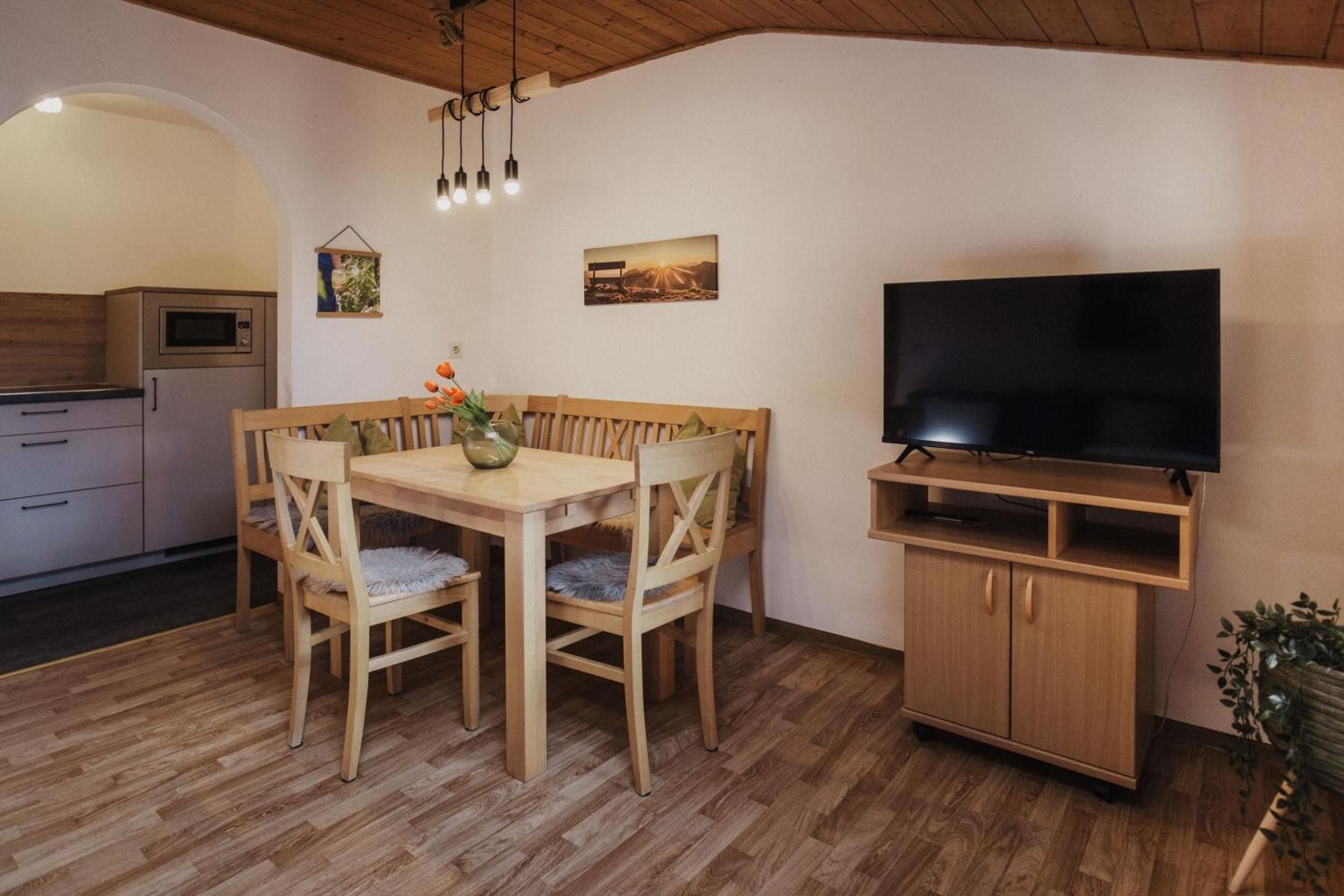 Un comedor con mesa y sillas de madera, un televisor sobre un mueble bajo y una cocina al fondo.