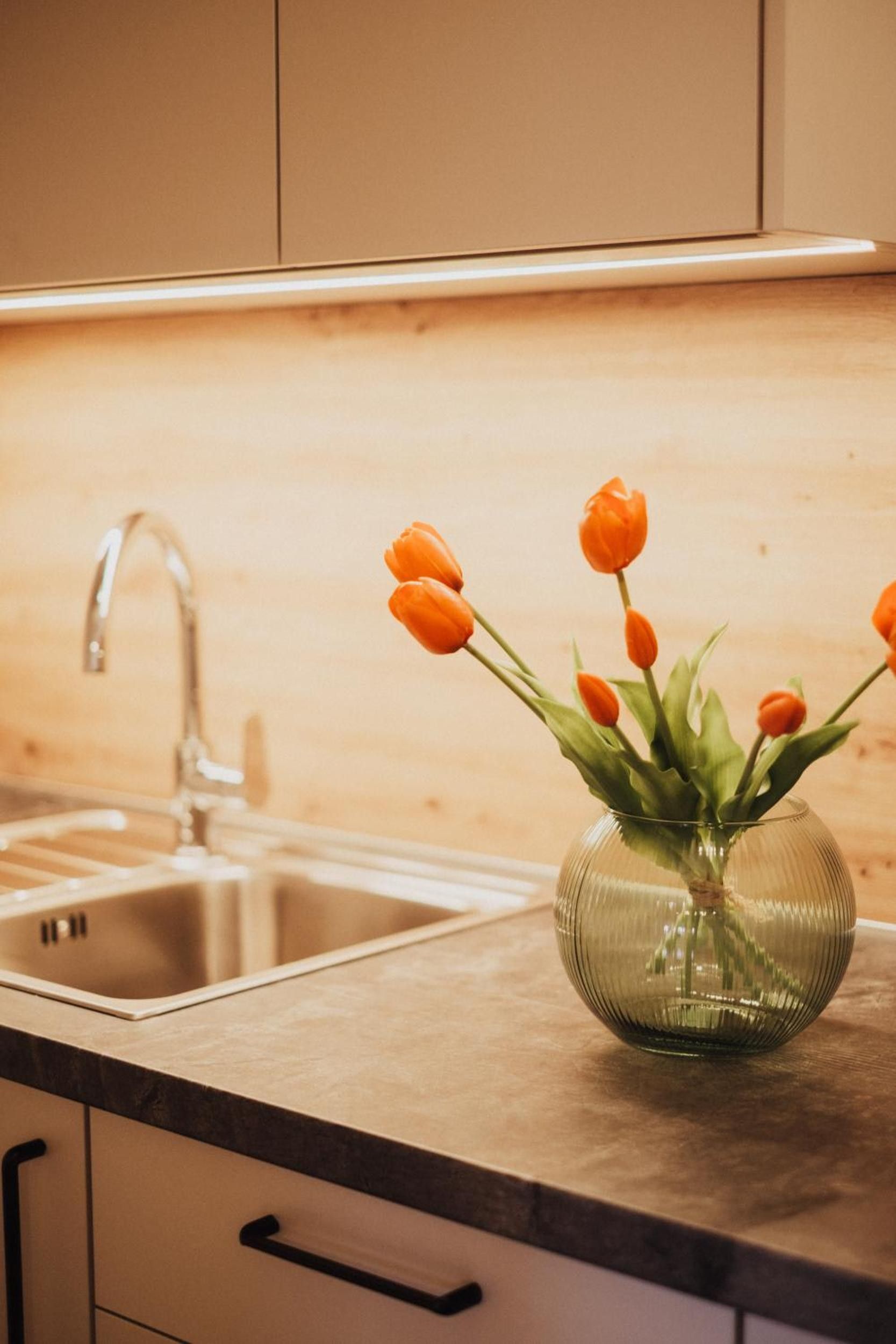 Une cuisine avec un évier en acier inoxydable et un vase en verre contenant des tulipes orange, posé sur le plan de travail.