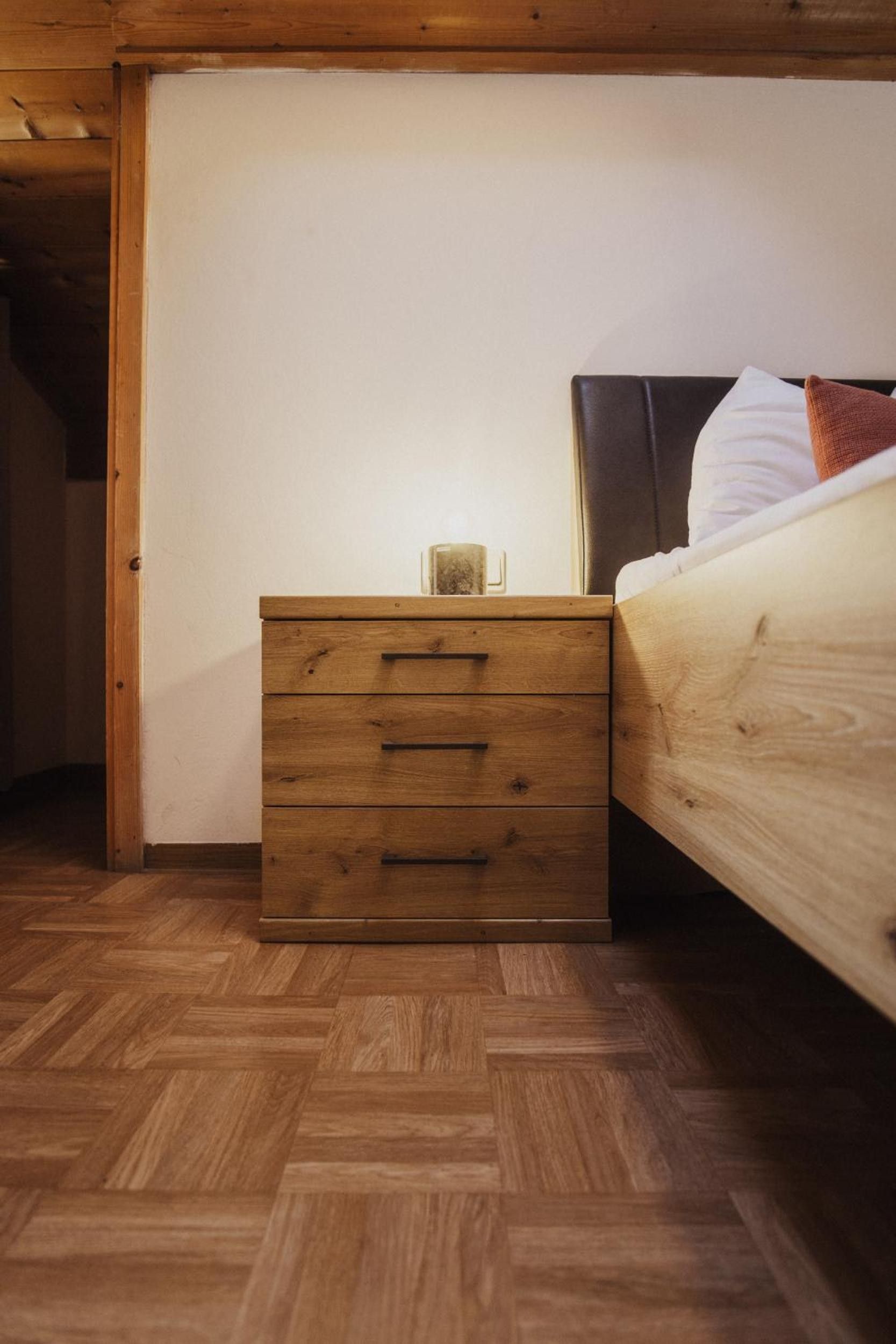 Una mesita de noche de madera con cajones y una lámpara de mesa pequeña, ubicada junto a una cama con cabecera y almohadas.