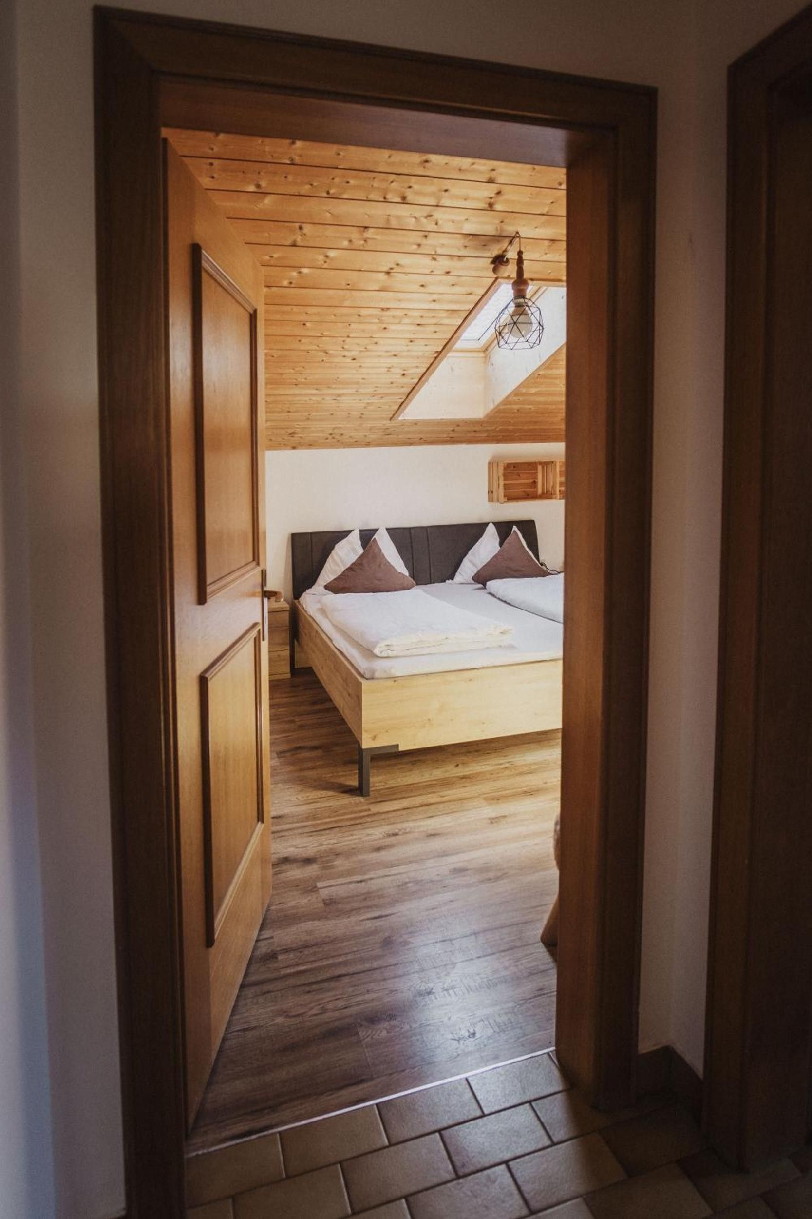 A bedroom with a wooden floor, a bed with a headboard, and a sloped ceiling with a hanging light fixture.
