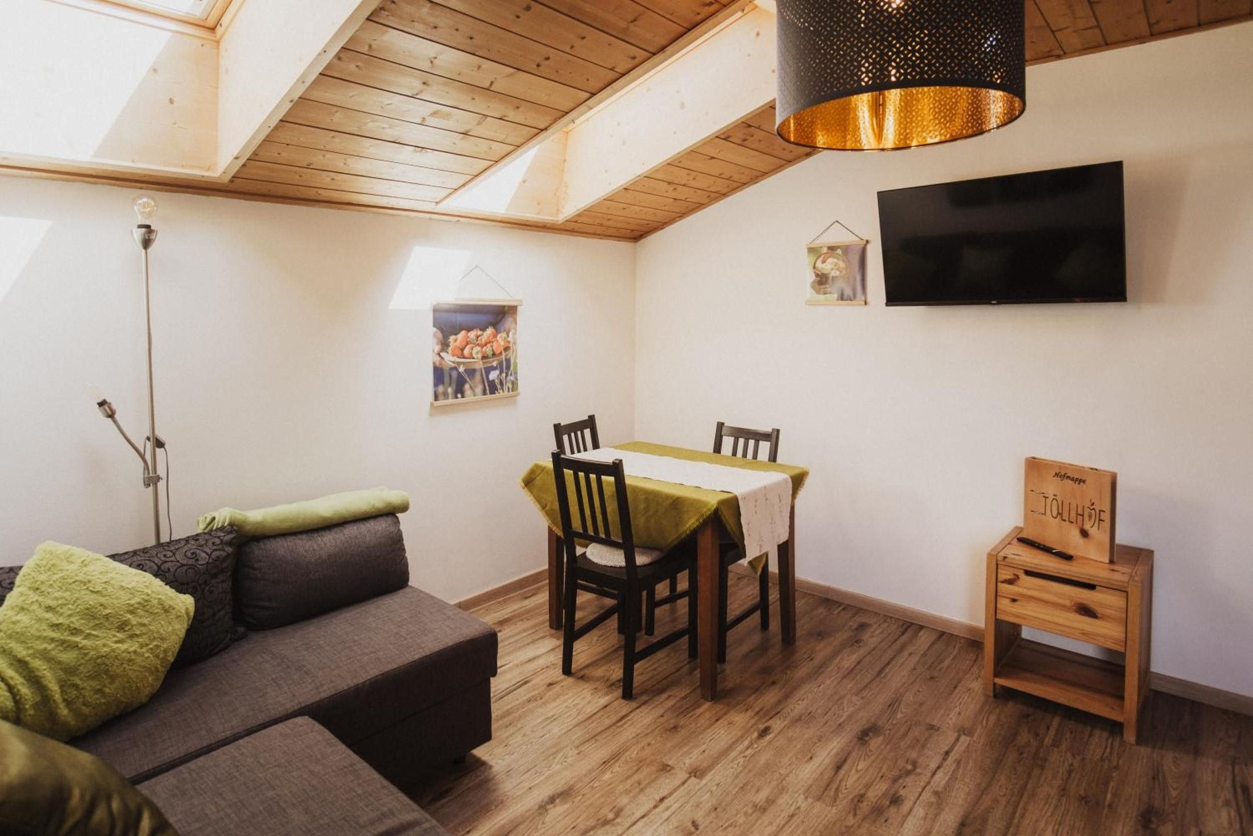 A cozy room with a wooden ceiling, a dining table with chairs, a sofa with cushions, a TV mounted on the wall, and a small wooden cabinet.