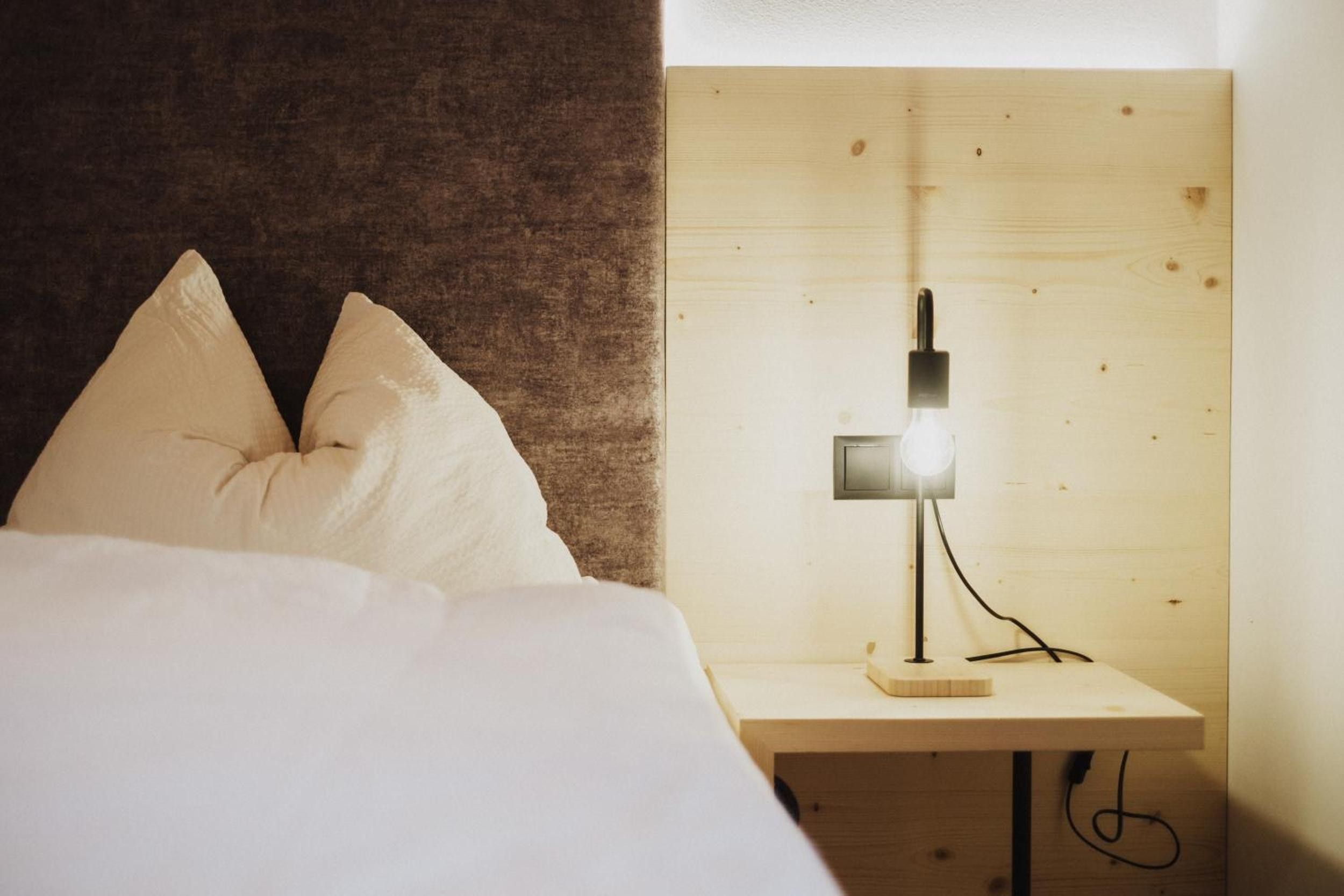 Una cama con ropa de cama blanca y dos almohadas, una mesita de noche con una lámpara y un cabecero de madera.