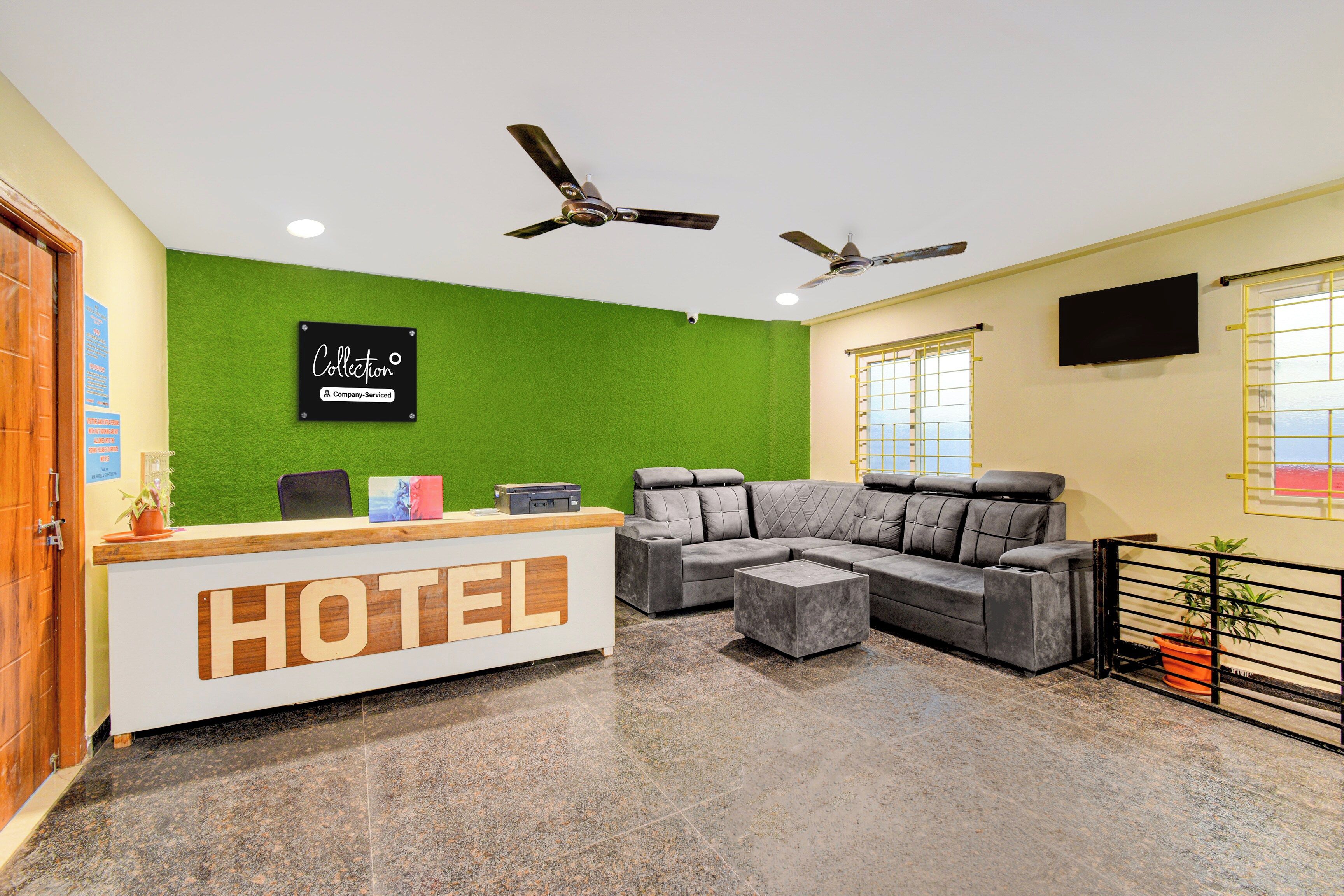 A hotel reception area with a reception desk, a grey sectional sofa, two ceiling fans, and a green wall.