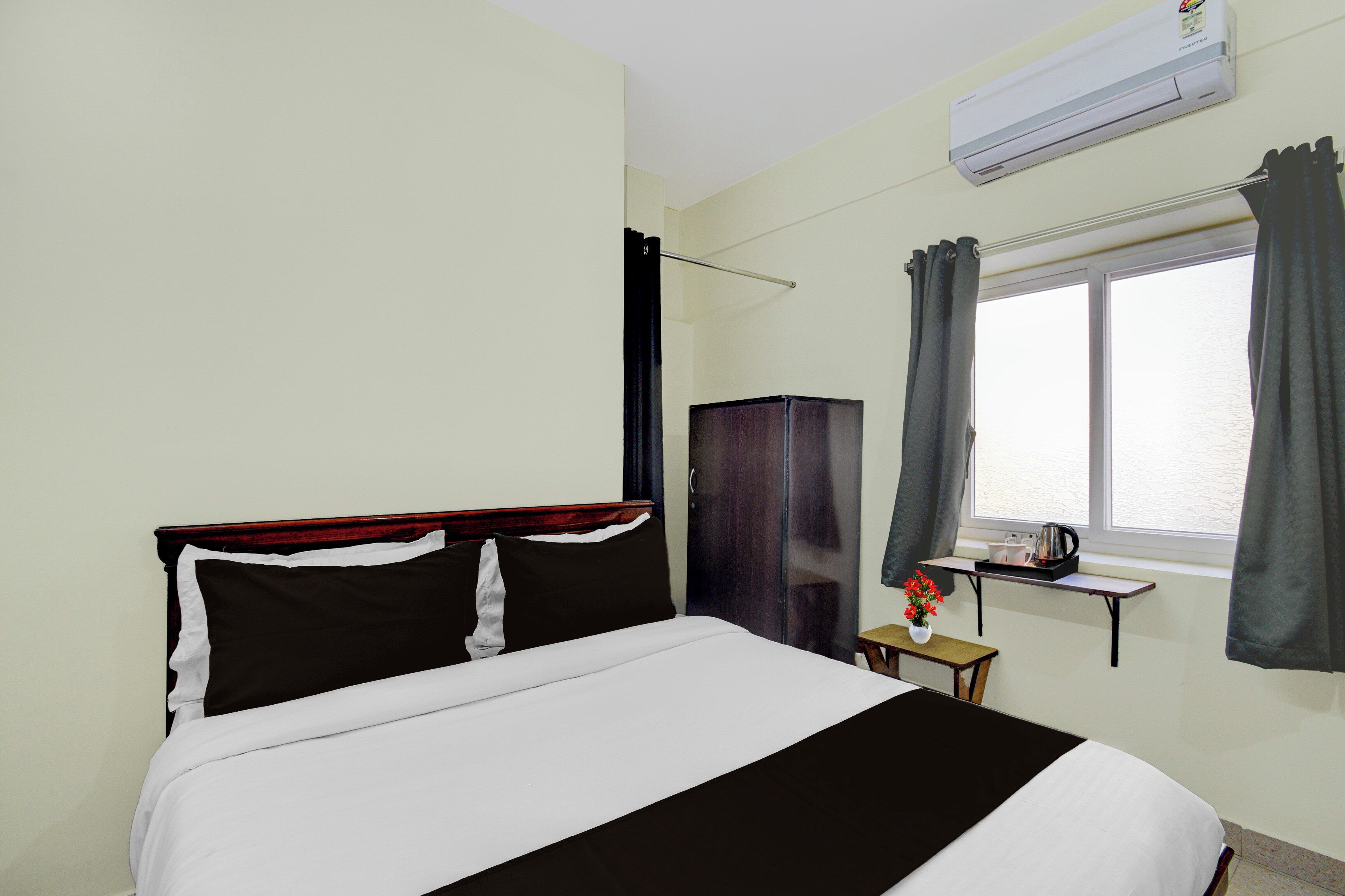 Una habitación de hotel con una cama, un armario, un ventanal con cortinas, una mesita con una tetera y un equipo de aire acondicionado.