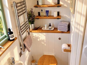 A compact bathroom with a towel rack, shelves with toiletries, a toilet, and a window. - Oak Hut (Bishops Castle)