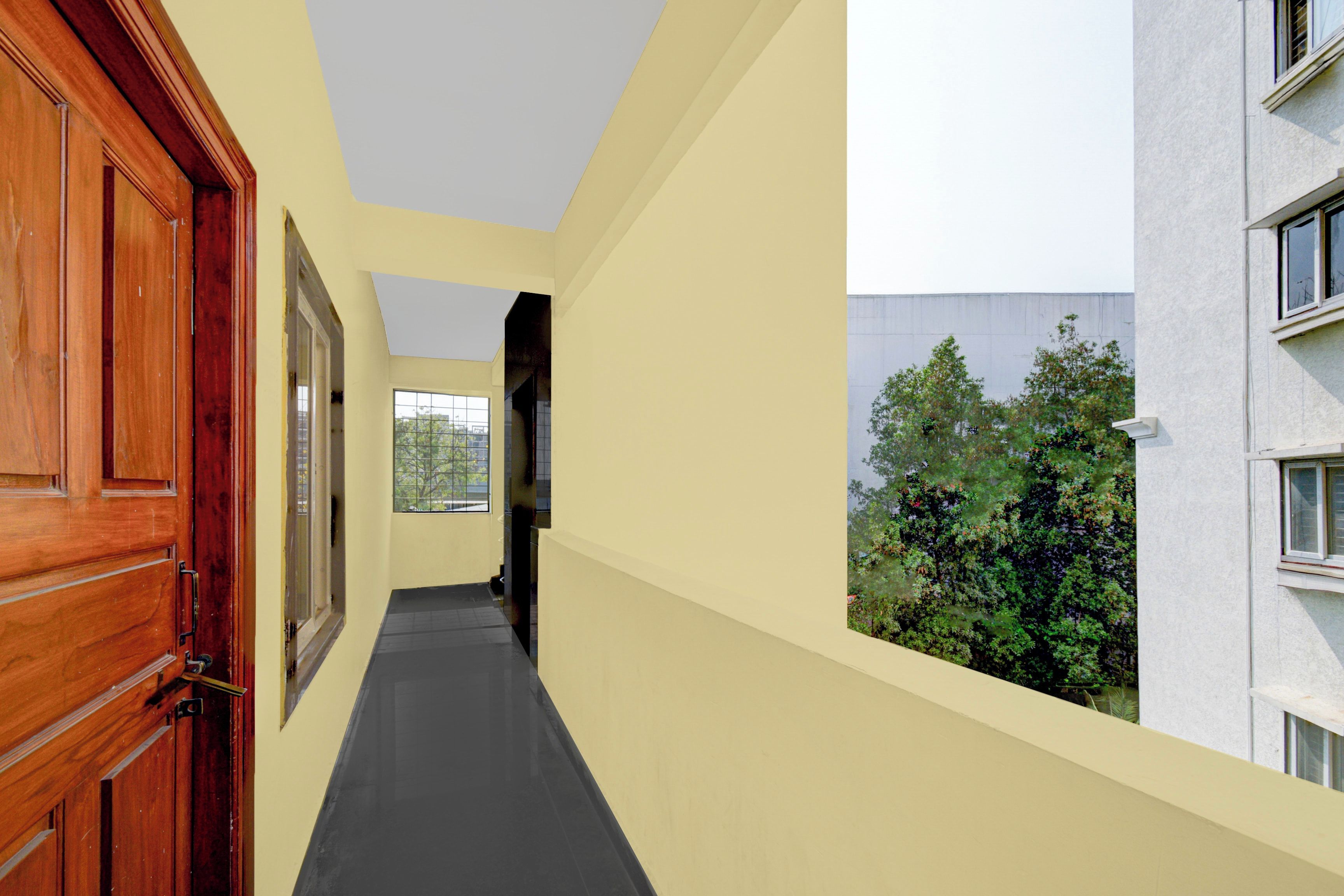 A narrow corridor with a wooden door on the left, a window with a view of trees, and a building with multiple windows on the right.
