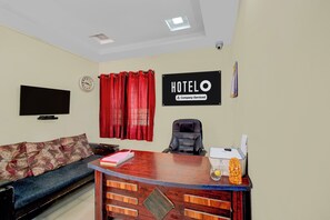 A hotel room with a wooden reception desk, a sofa, a TV, and a sign that reads 'HOTEL O Company Serviced'.